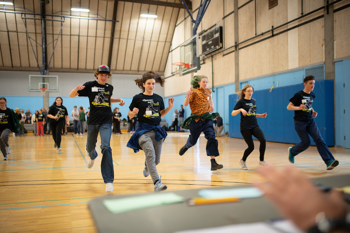 Male and female high school students sprinting across gym to reach desk with pencils and papers.