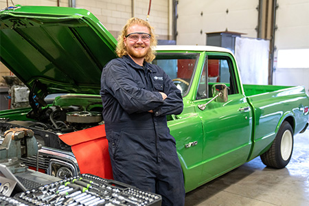 An automotive student smiling and leaning back on a green truck with its hood open..