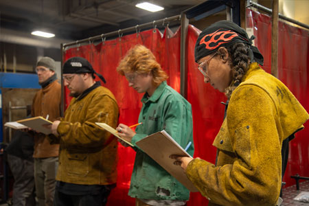 A group of students in a welding workshop, wearing protective jackets and headgear, standing in front of red welding curtains while writing notes on clipboards.