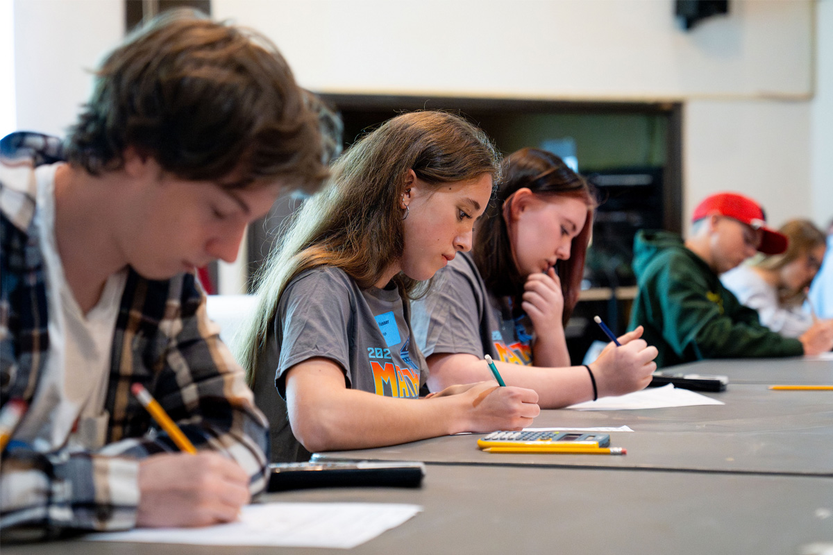 A row of students sits at a long table, writing on papers during a math contest exam round.
