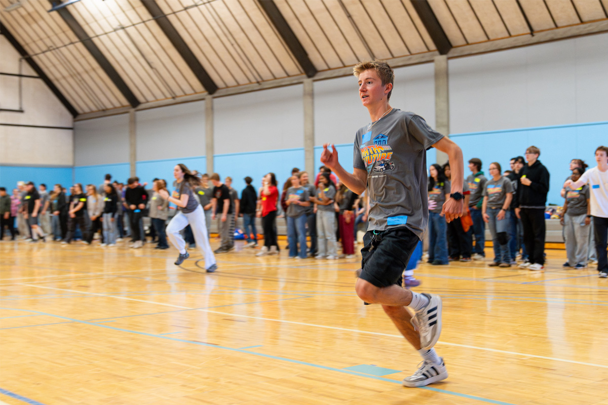 A student runs across a gymnasium floor during a math contest activity, with a large group of students watching from the sidelines.