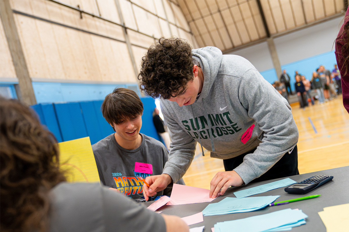 Two students work together at a table covered with colored paper and a calculator in a gymnasium during the COCC Math Contest.