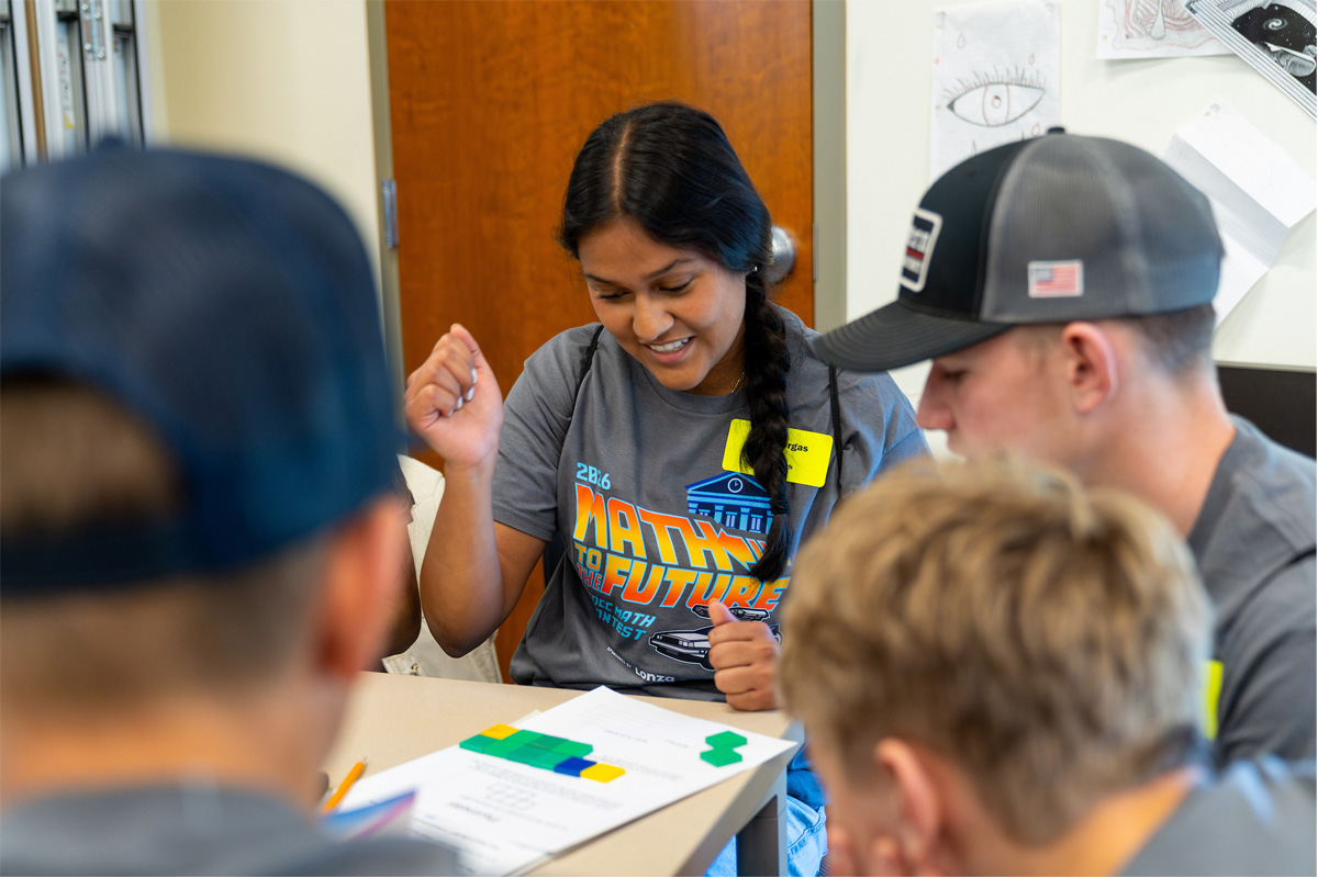 A student smiles and gestures enthusiastically while working on a puzzle activity with a small group of students at a table.
