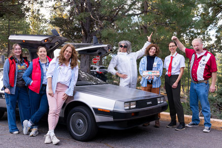 A group of seven people dressed in Back to the Future costumes pose beside a silver DeLorean with its gull-wing doors open, surrounded by pine trees.