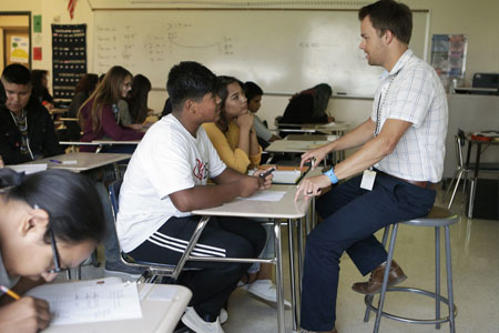 A math instructor leans in to help a student while others work at their desks.